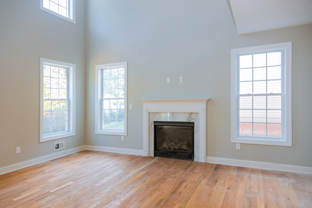 Empty living room with newly installed hardwood flooring, light gray walls, three windows and a fireplace
