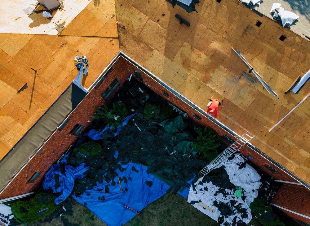 Overhead view of workers stripping old roof sheathing on a brick building with tarps below for debris – Roof Replacement