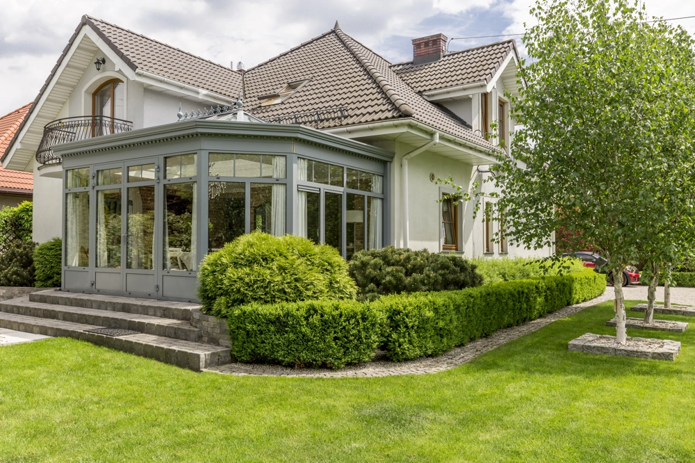 Front exterior room addition with glass-enclosed sunroom, new roof and landscaped shrubs
