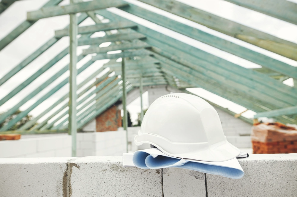 Hard hat and blueprints on a block wall under green roof trusses during Home Remodeling