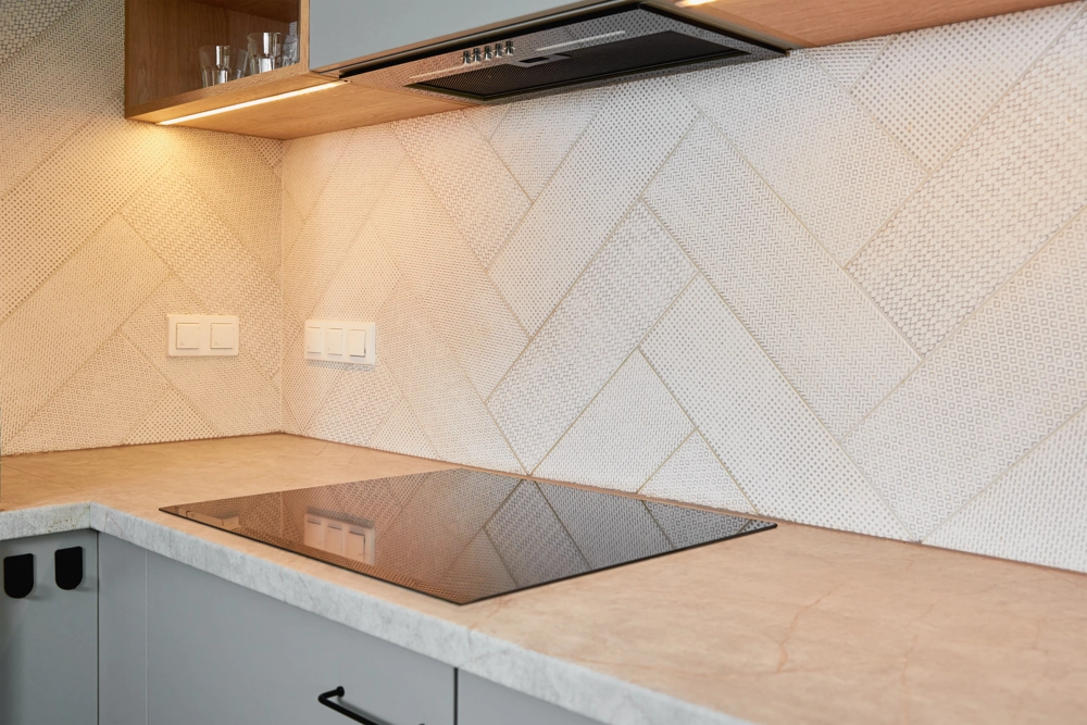 Modern kitchen backsplash tile installation above induction cooktop on stone countertop under cabinet lighting